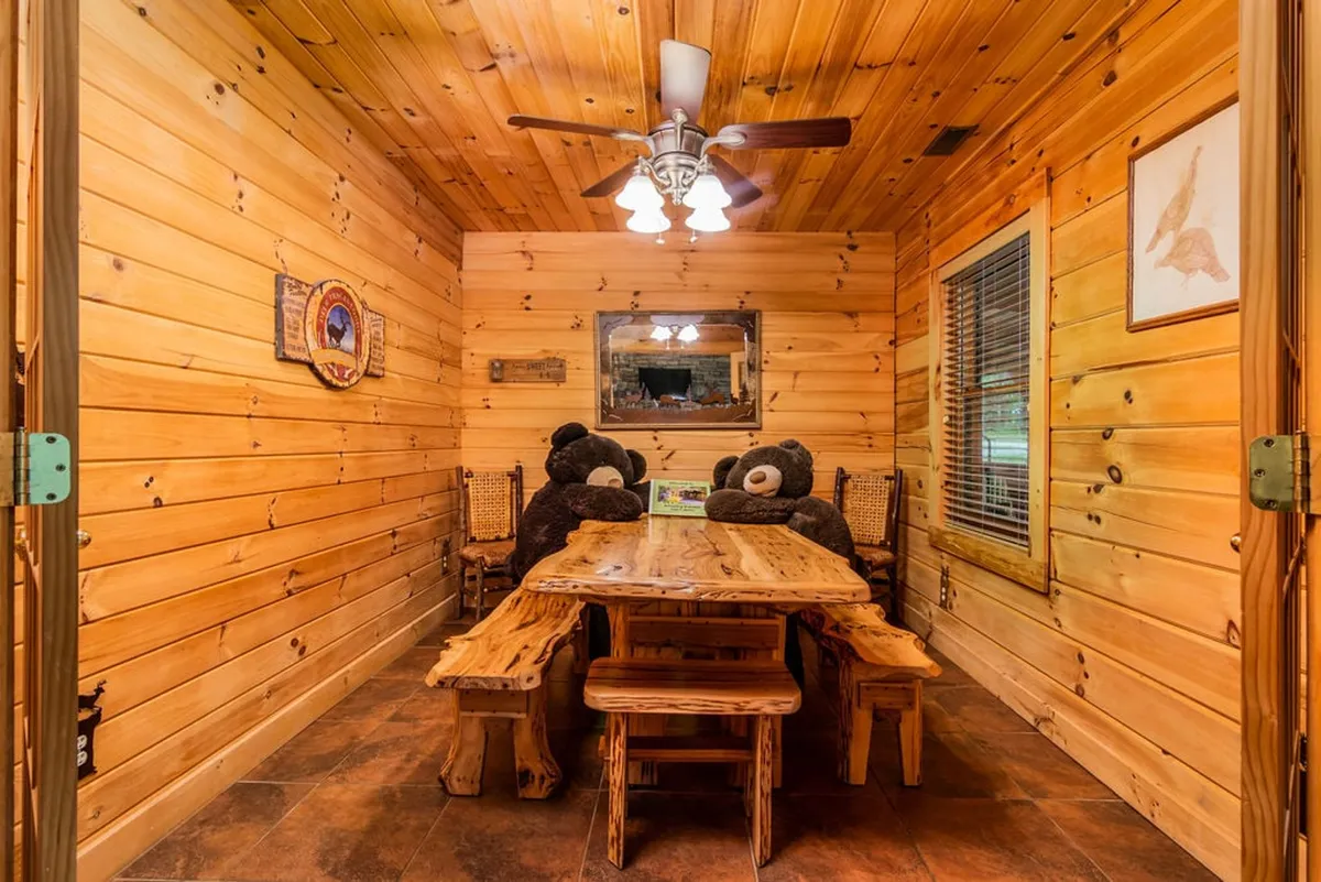 A cozy dining room featuring a wooden table surrounded by comfortable chairs, wood-paneled walls, a ceiling fan, and stuffed bears that add a whimsical, family-friendly touch. The space is clearly designed for family dinners, heartfelt conversations, and making memories together.