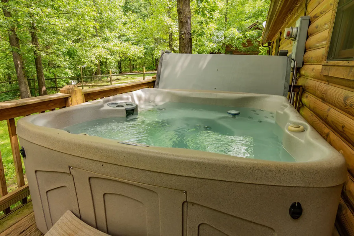 This exterior image showcases a private wooden deck with a hot tub surrounded by lush greenery, inviting guests to soak and unwind in a tranquil natural setting. The deck exudes a sense of seclusion, perfect for relaxing after a day of outdoor activities, with open sky views ideal for stargazing.