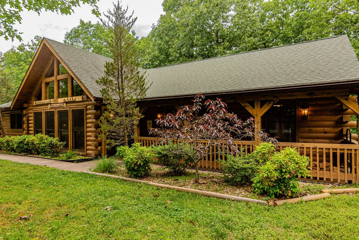 This image showcases the log cabin structure of Deer Tracks Lodge, its inviting wooden porch, and the landscaped garden area. The open outdoor space is visible, suggesting plenty of room for pets to roam. The porch with seating is ideal for relaxing while watching pets play safely nearby.