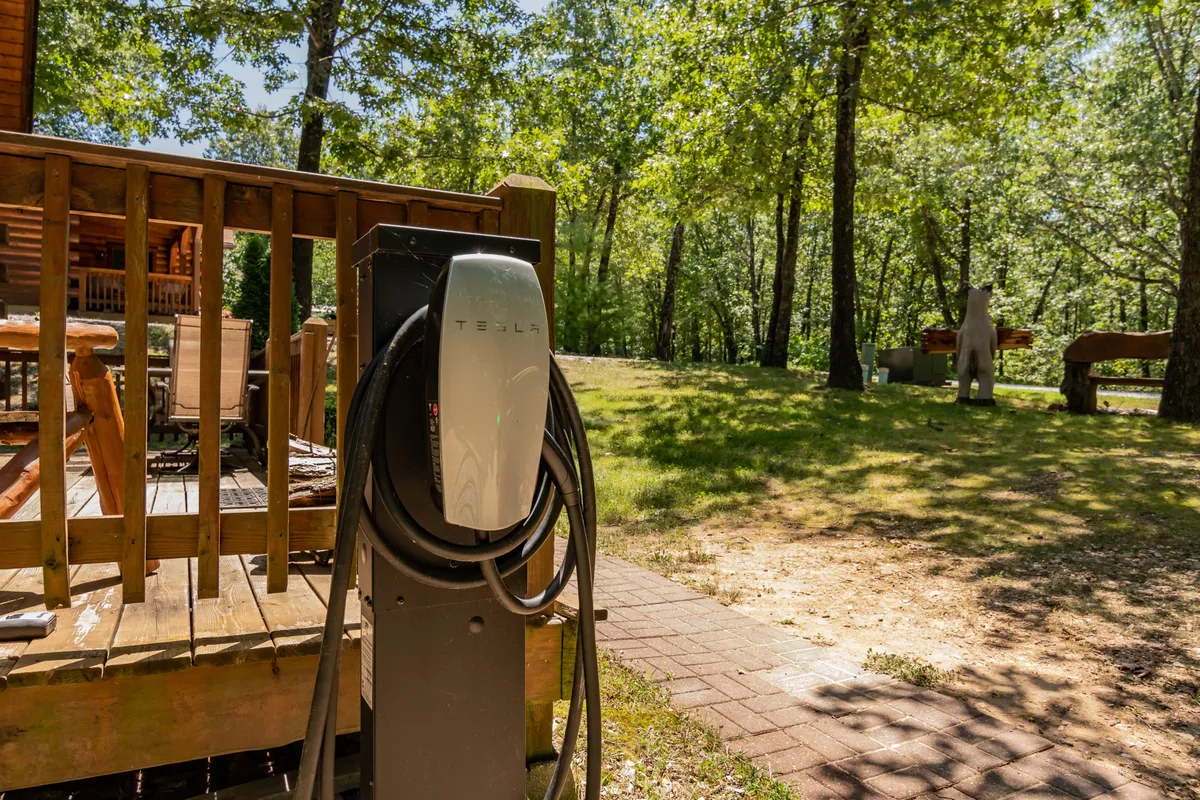 This image focuses on the Tesla charging station situated near the wooden deck, with surrounding trees and outdoor seating visible. The area exudes a relaxed, eco-friendly vibe, emphasizing modern convenience nestled in nature.
