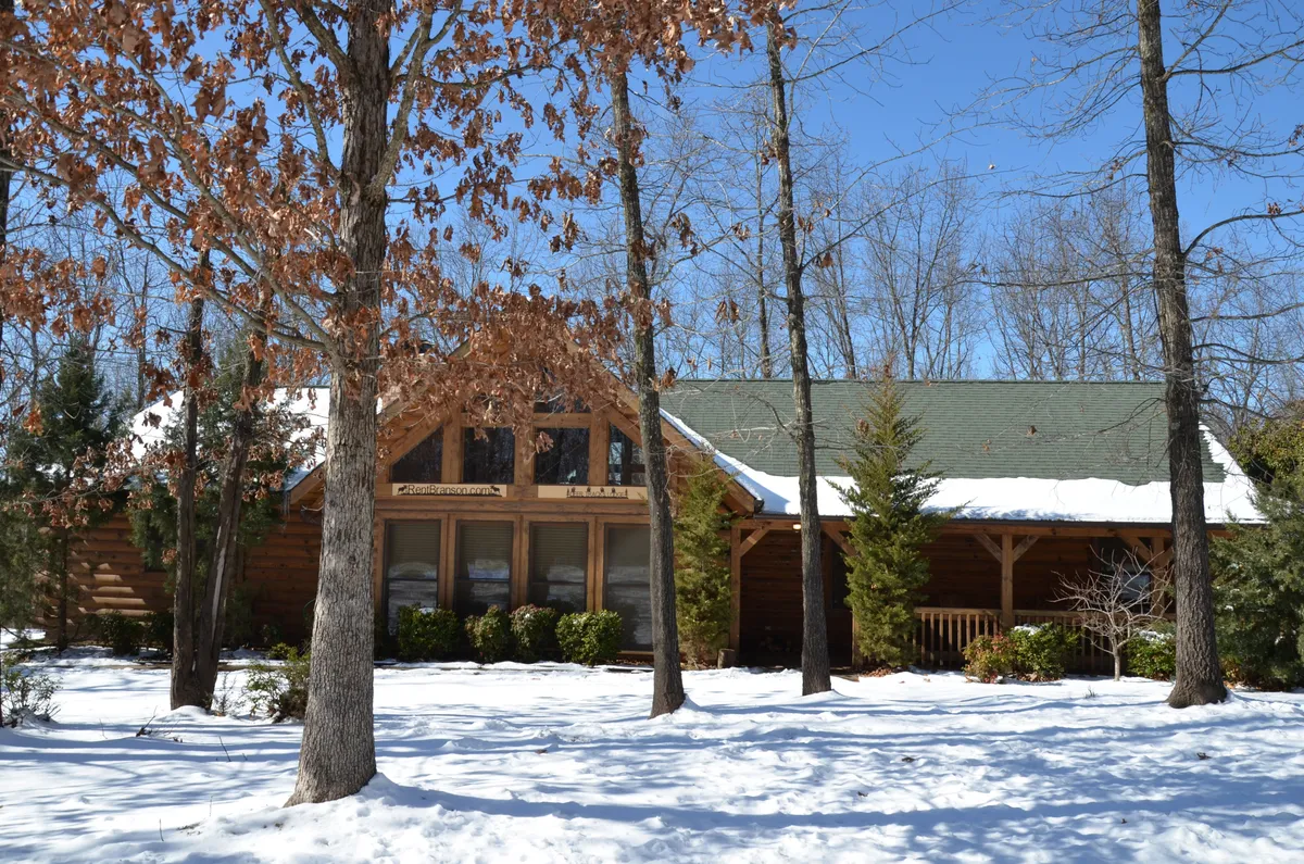 This exterior shot captures the log cabin structure of Deer Tracks Lodge surrounded by trees and a light layer of snow. Large windows are visible, and the cabin sits amidst a serene, natural setting that hints at tranquility and proximity to outdoor activities.