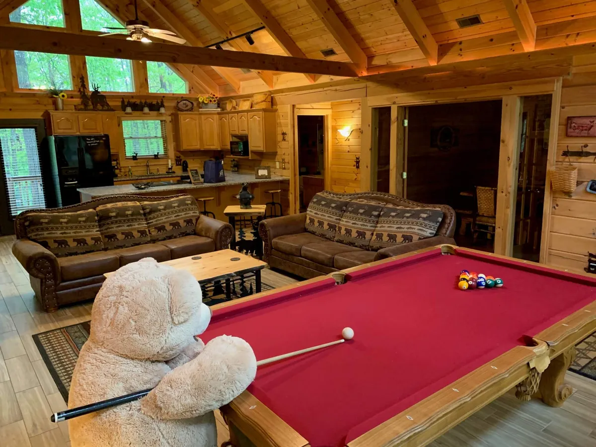 A cozy, playful interior featuring a pool table, large teddy bear, comfortable sofas, and an open kitchen area. The space radiates warmth and is clearly designed for fun family time and relaxation after a day of exploring.