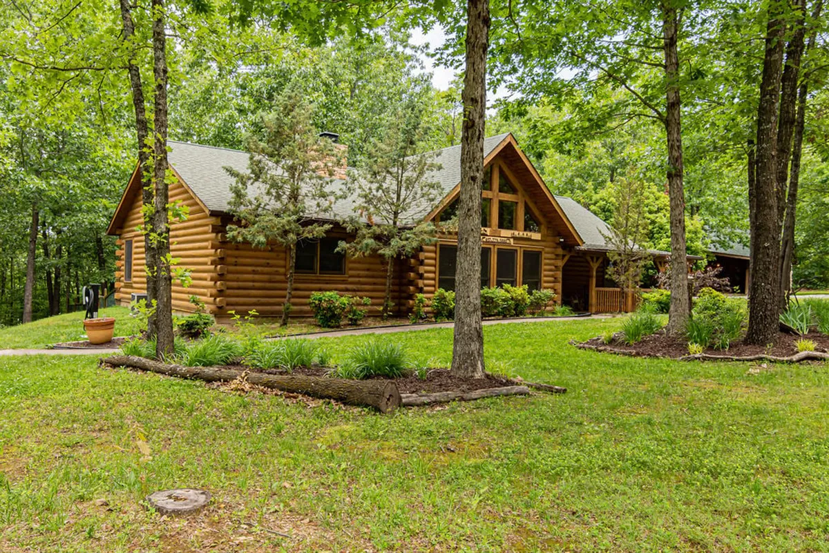 A cozy log cabin exterior with large front windows, green landscaping, and mature trees in the background. The scene evokes feelings of peaceful seclusion and a strong connection to nature.