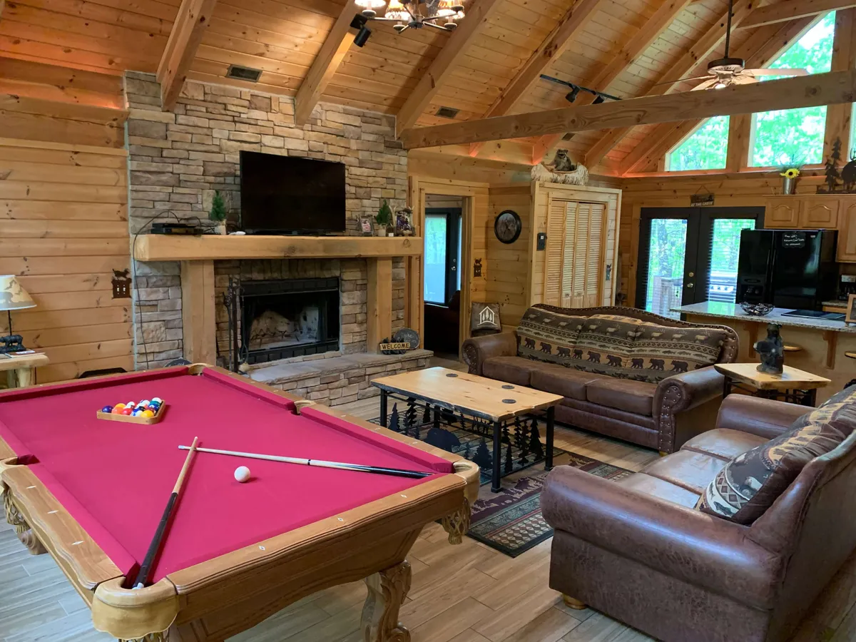 A warm, friendly family room scene showcasing a billiards table positioned near a crackling fireplace, a comfortable sofa set, exposed wooden beams, and a mounted TV. The setup suggests a space for relaxing, socializing, and enjoying game nights together.