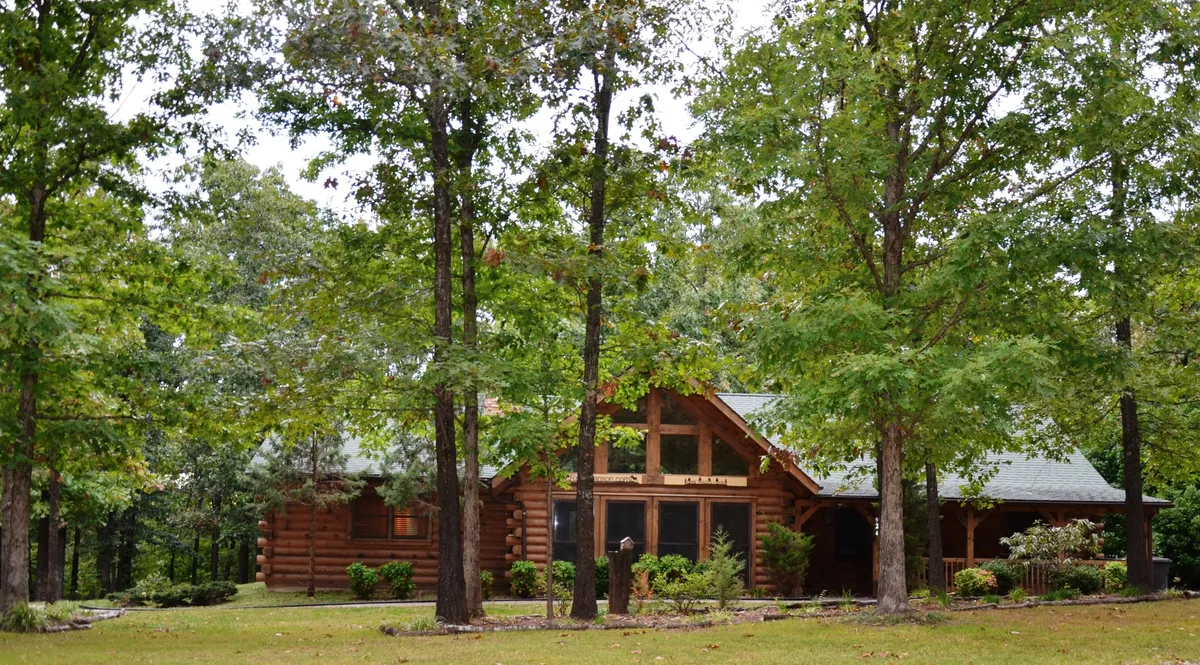 A welcoming log cabin exterior nestled among lush green foliage, featuring a spacious front porch with outdoor seating. The inviting atmosphere suggests a serene escape in nature while maintaining accessibility to the nearby community.