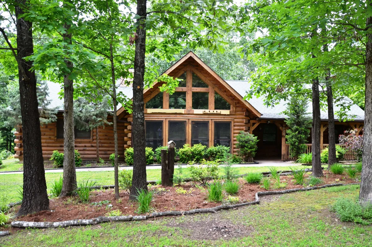The exterior of a rustic log cabin with large front windows and lush greenery all around. The porch invites gatherings or quiet moments, and the surrounding landscape hints at barbecue evenings and nature walks, resonating with the section's themes of socializing and connecting in the Ozarks.