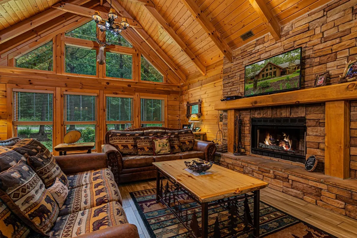 The hero image features a warm, inviting living room with soaring wooden beams, a majestic stone fireplace, expansive windows overlooking the Ozarks, and plush couches arranged for cozy gatherings.