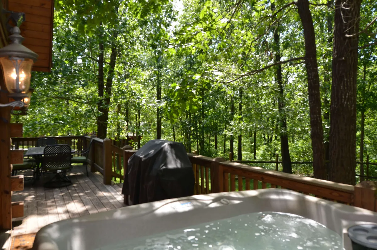 This image features a spacious wooden deck surrounded by lush trees, equipped with a bubbling hot tub, a propane grill, and comfortable outdoor furniture. The setting conveys a tranquil, private retreat perfect for outdoor relaxation and dining.