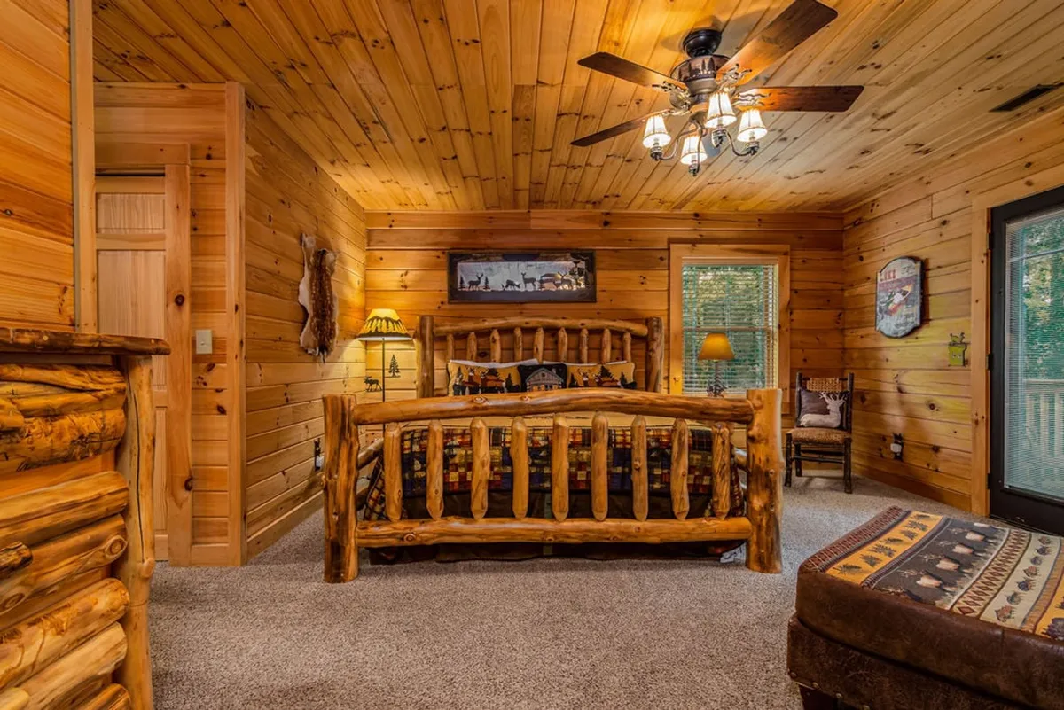 A warm and welcoming bedroom featuring a rustic log bed, wooden ceilings, a chandelier, decorative artwork, and a window view that lets in natural light. The bed is dressed in plush linens, creating a peaceful and cozy retreat.