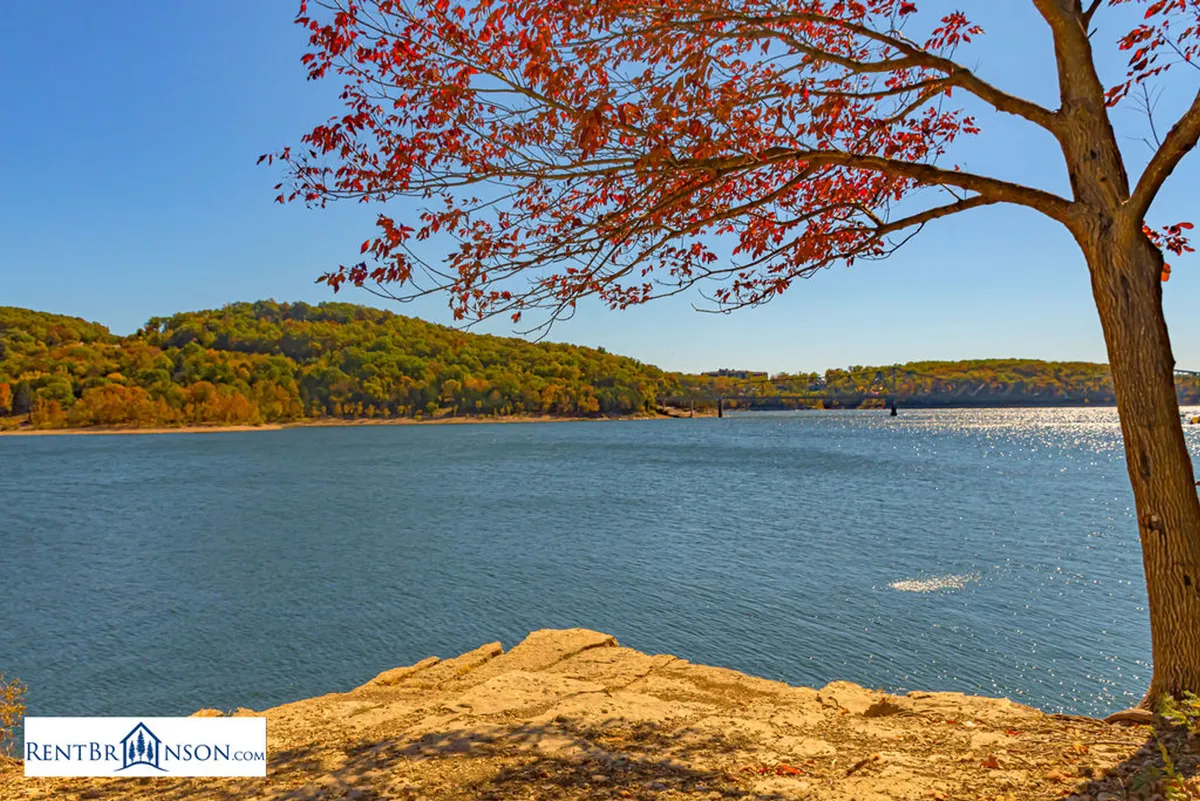 A serene water view with autumn leaves, a rocky shore, and hills in the background. The peaceful scene invites thoughts of fishing, boating, and hiking—activities synonymous with exploring the Ozarks' natural beauty.