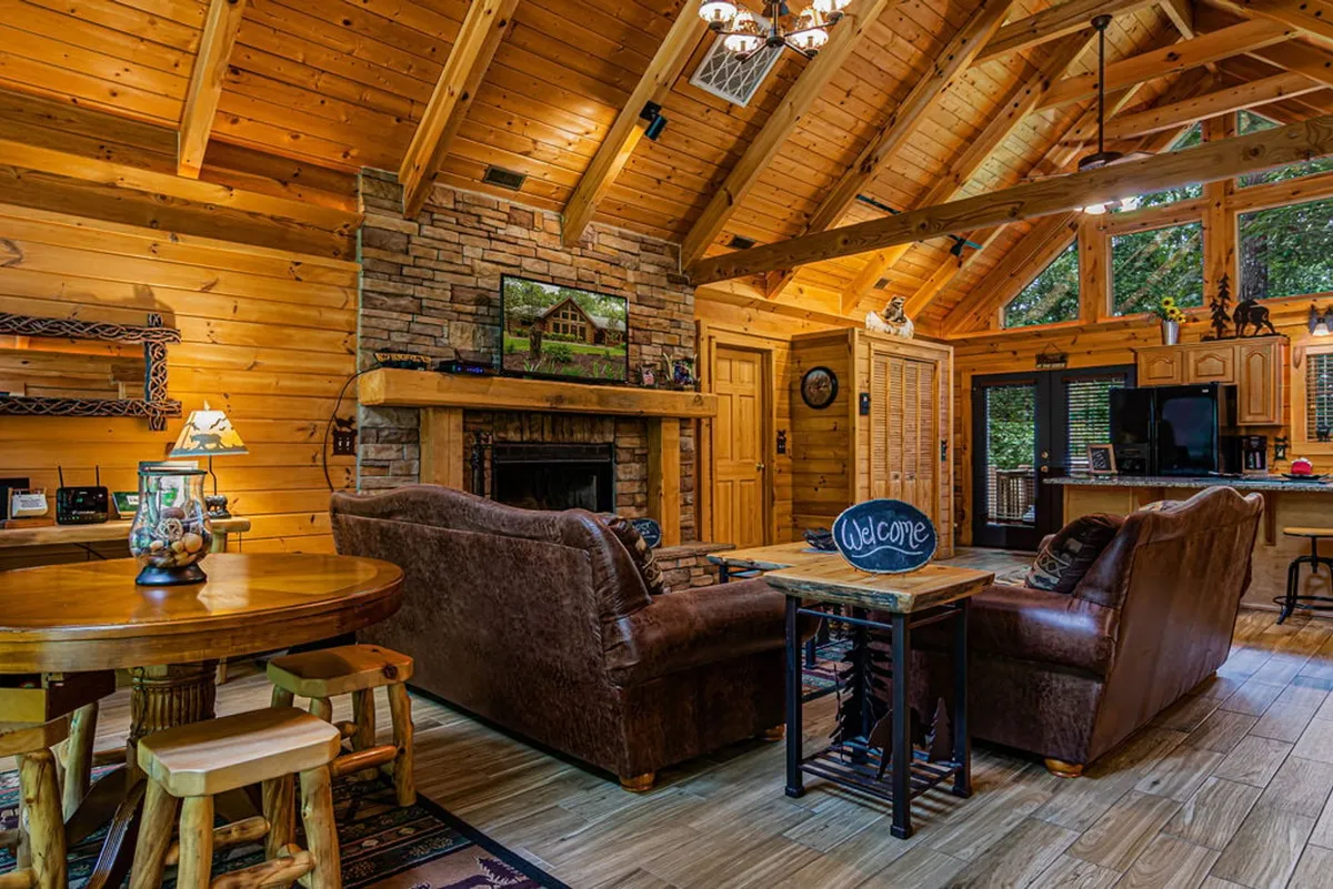 This image captures an expansive open-concept living area with a prominent wood-burning fireplace as the focal point, complemented by rustic wooden beams overhead. The space seamlessly flows into the dining area and kitchen, featuring comfortable lounge seating and a large dining table—ideal for gathering. The kitchen area is visible in the background, tying together the communal feel.