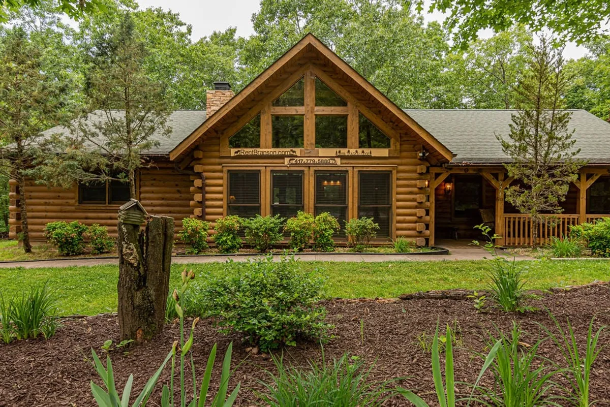 A warm, welcoming log cabin exterior with large front windows and a landscaped garden. The porch offers space for gathering, and the natural surroundings provide a sense of adventure and relaxation, ideal for families preparing to set out on nearby explorations.