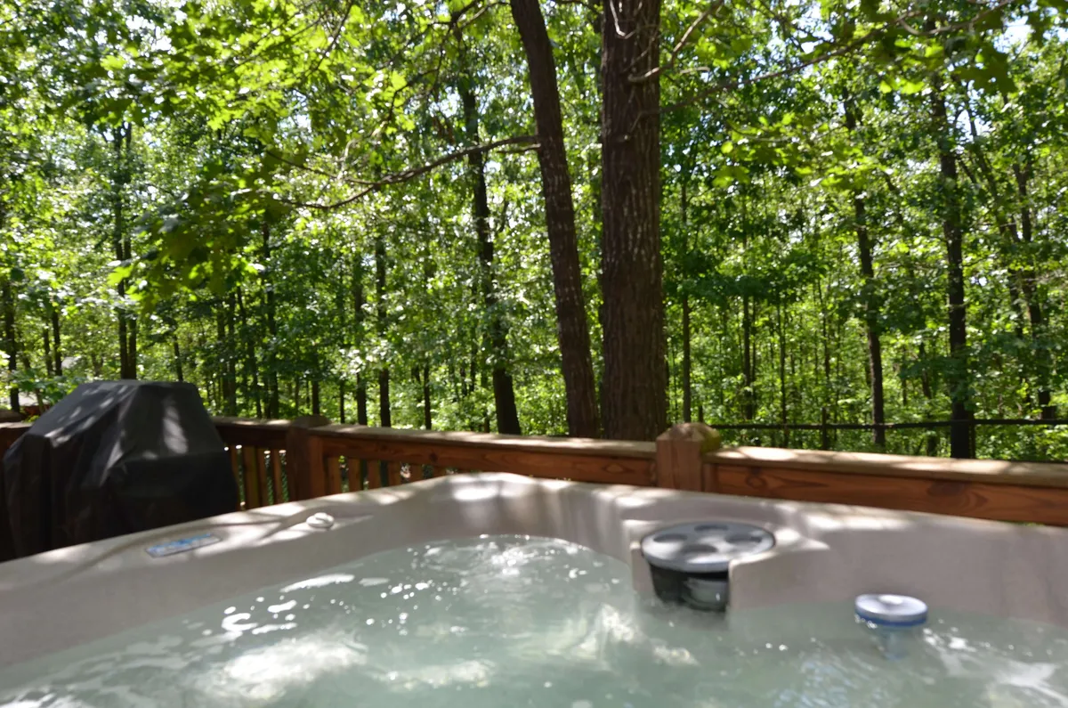 The image shows a deck area with a hot tub overlooking a wooded landscape. The deck is framed by a railing, and tall trees provide privacy and a sense of seclusion, while the hot tub suggests relaxation after a busy day.