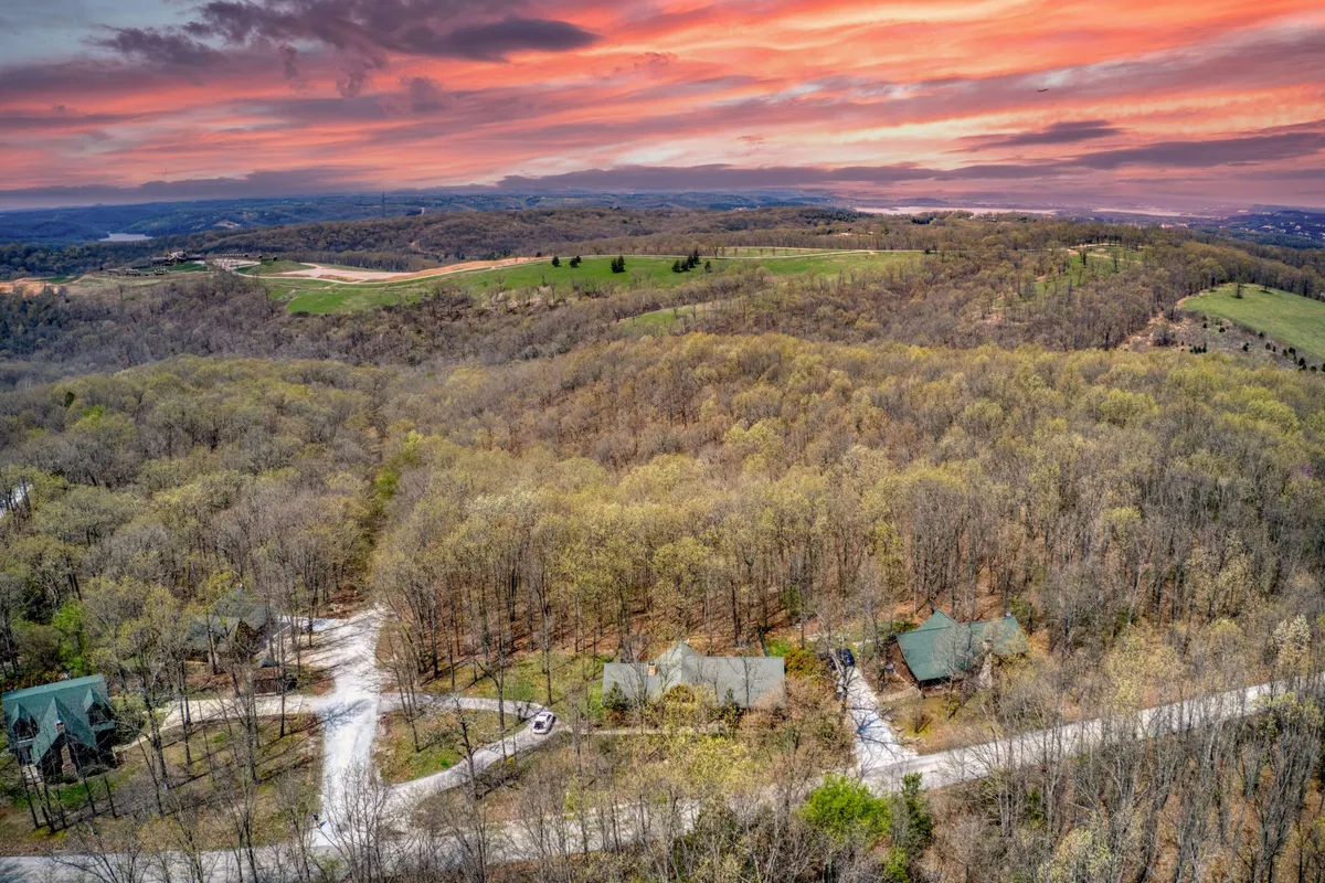 A sweeping view of the Ozarks landscape with a scenic tree canopy, visible cabins, and winding roads, capturing the expansive outdoor environment around the property. The horizon glows with sunset colors, evoking a peaceful, adventurous spirit ideal for hiking, exploring, or gathering outdoors.